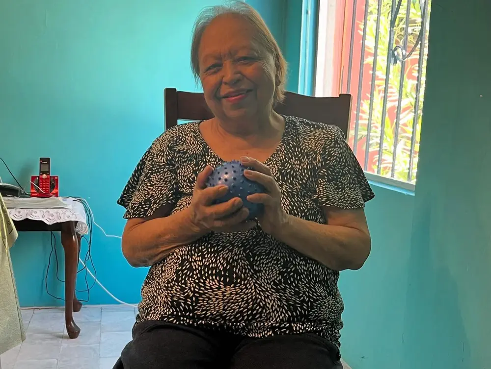 Una señora mayor sonríe mientras sostiene una pelota sensorial azul, sentada en casa.