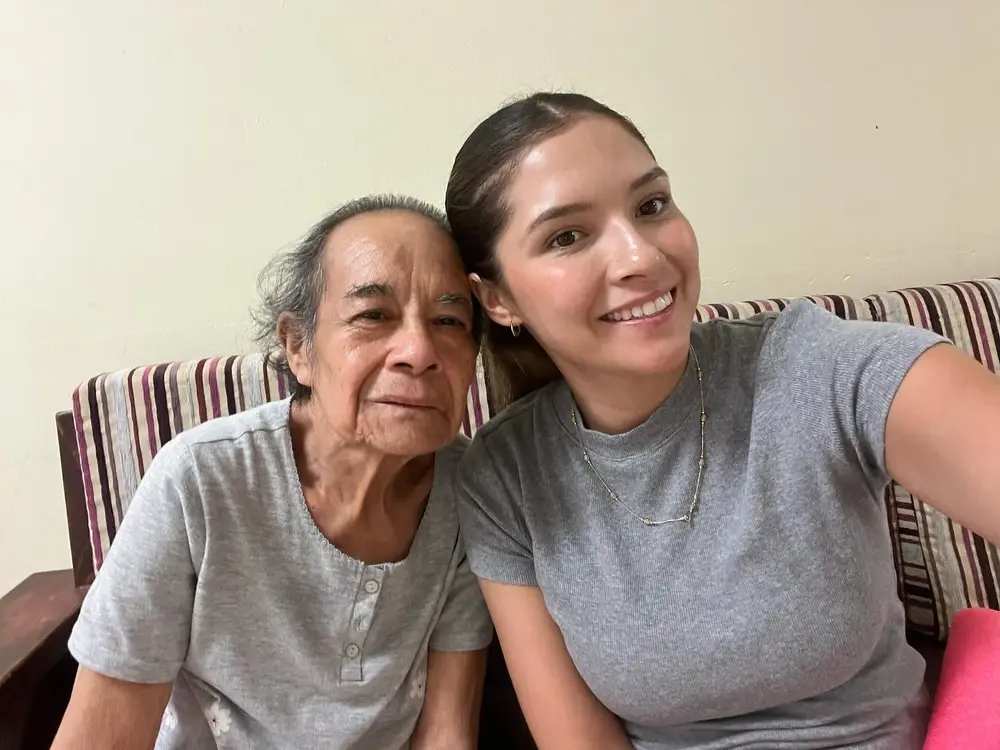 Karla, mejilla con mejilla, junto a una persona mayor a la que acompaña; ambas sonríen.
